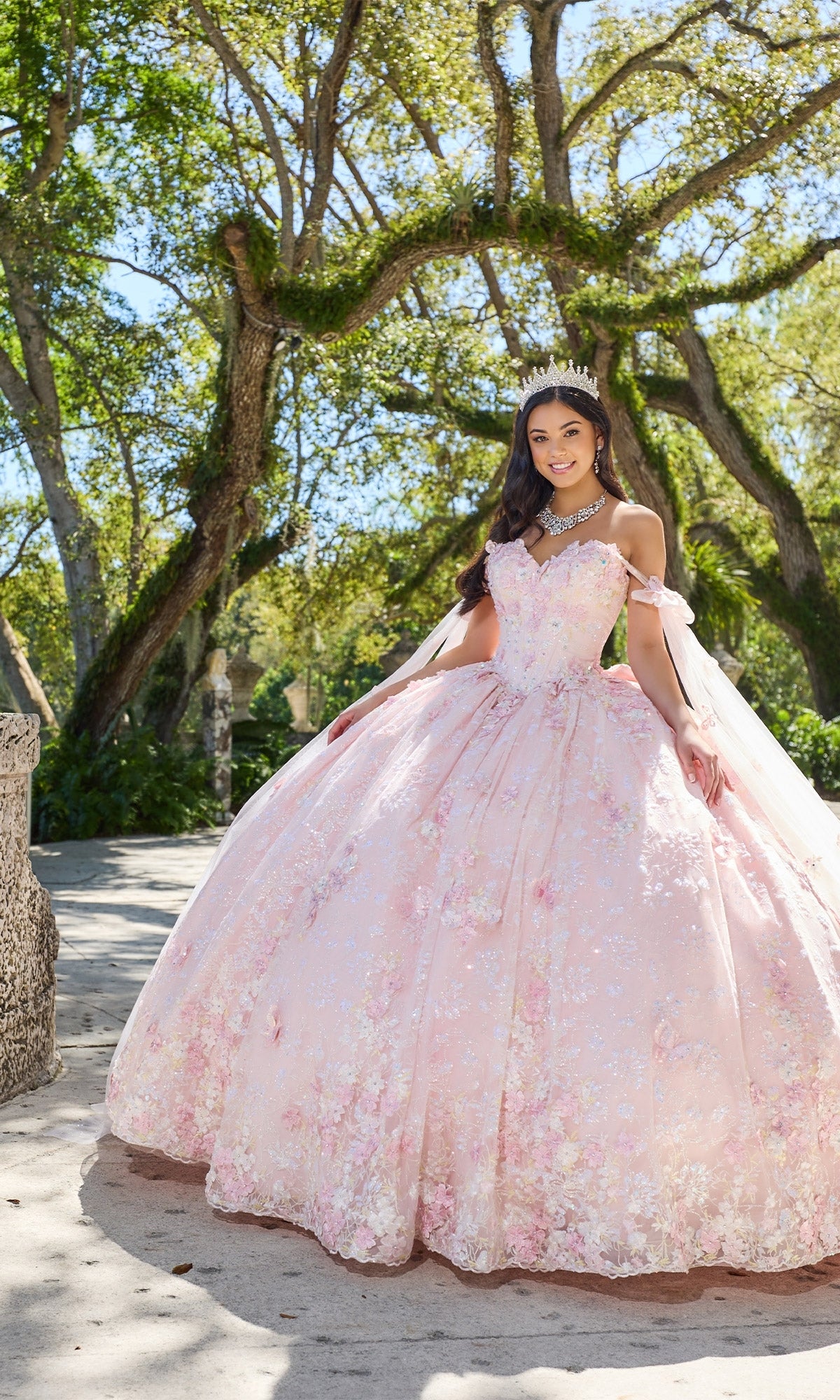 A young woman stands outdoors in the Princesa PR30135 3D Floral Quinceanera Dress with a corset bodice and tiara, surrounded by trees on a sunny day.